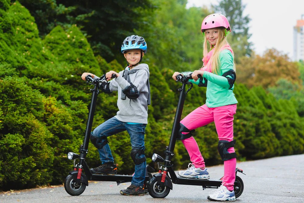 Två barn med hjälm, knä- och armbågsskydd står med var sin elsparkcykel på en asfalterad parkväg