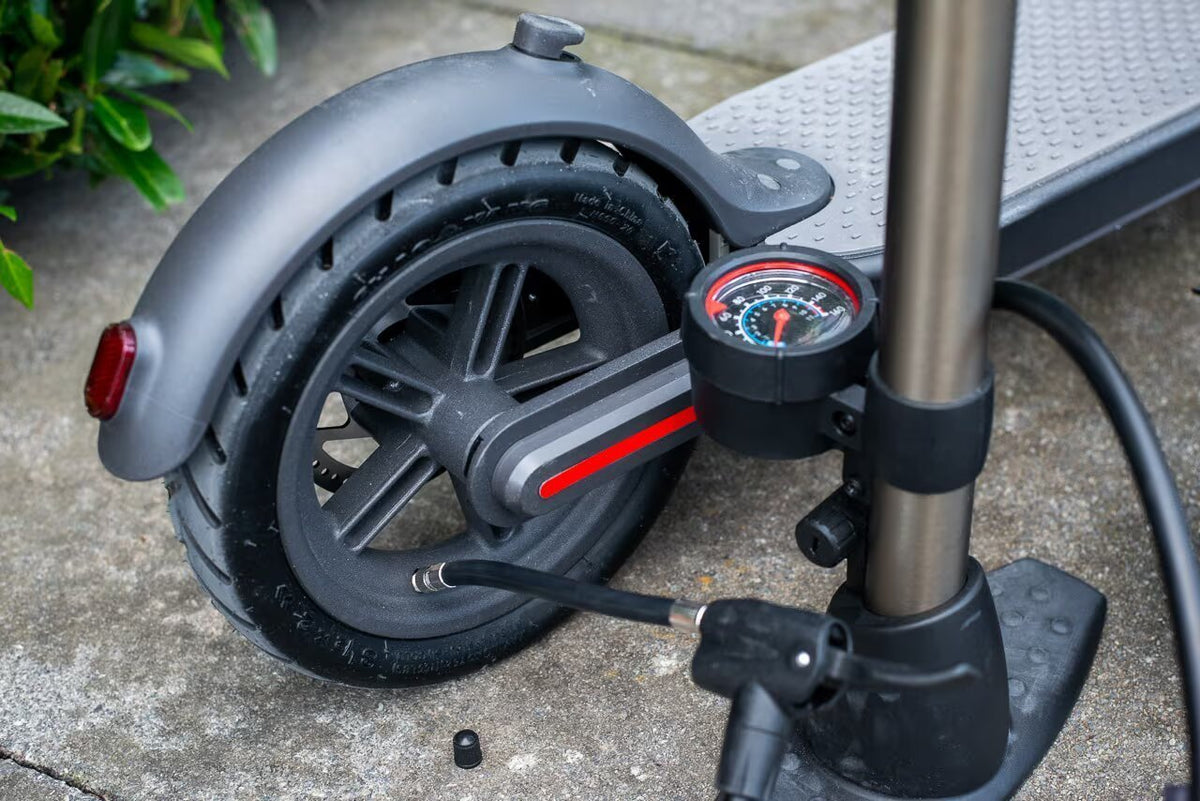 Standpump med tryckmätare kopplad till ventilen på bakhjulet till en mörkgrå elsparkcykel, ventilhatten ligger på marken