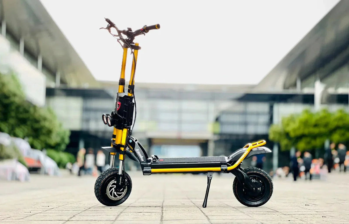 Gul och svart Kaabo-elsparkcykel med dubbelt styrrör, två strålkastare och grova terrängdäck, parkerad på ett stenlagt torg