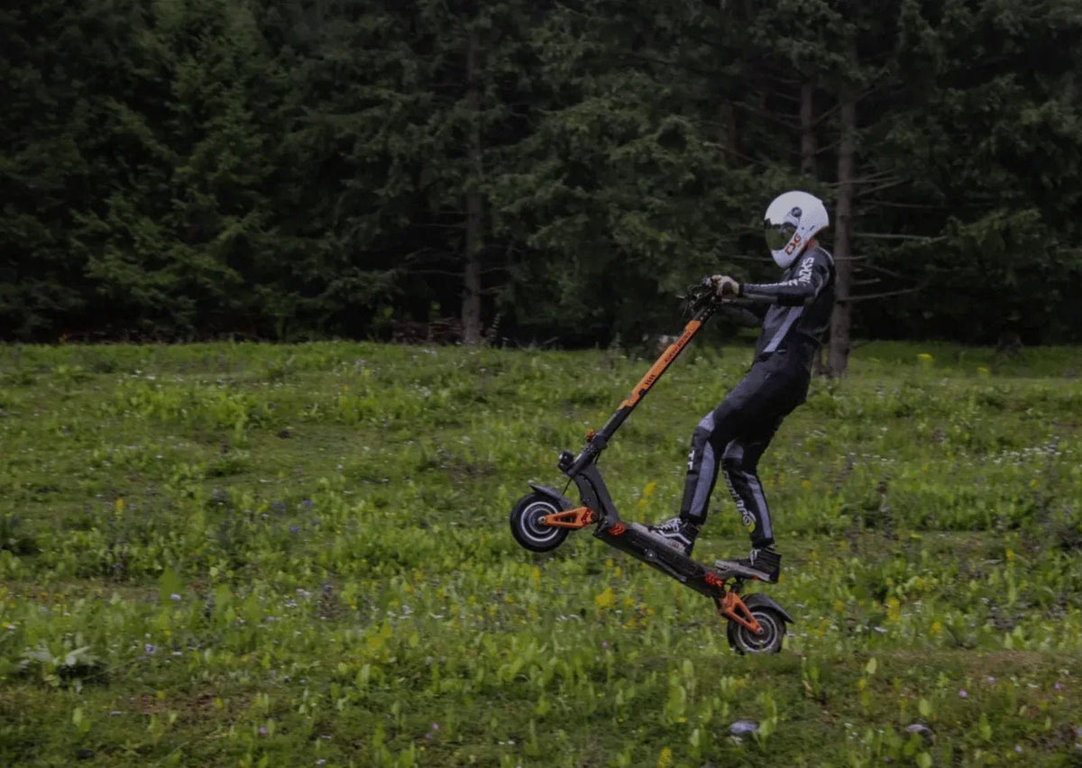 Förare i vit hjälm och skyddsdräkt hoppar med en svart elsparkcykel med orange detaljer över en blomsteräng framför granskog