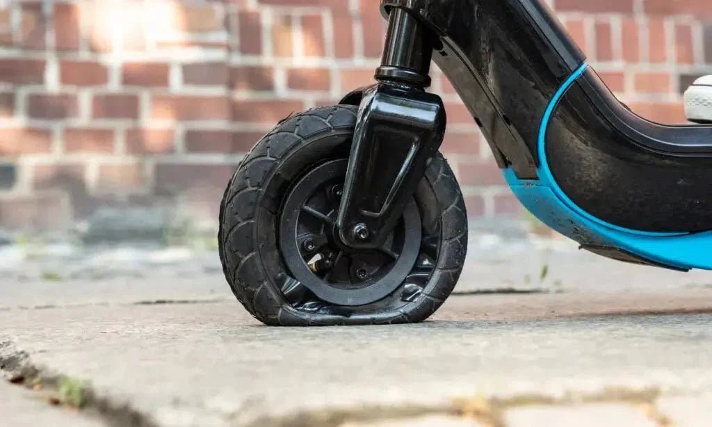 Helt platt luftdäck på framhjulet till en elsparkcykel, parkerad på en stenlagd gångväg framför en tegelvägg