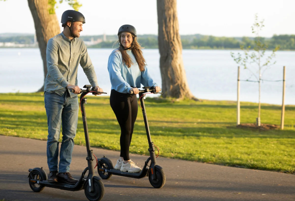 Två personer med hjälm kör NAVEE-elsparkcyklar på en asfalterad gångväg vid en sjö