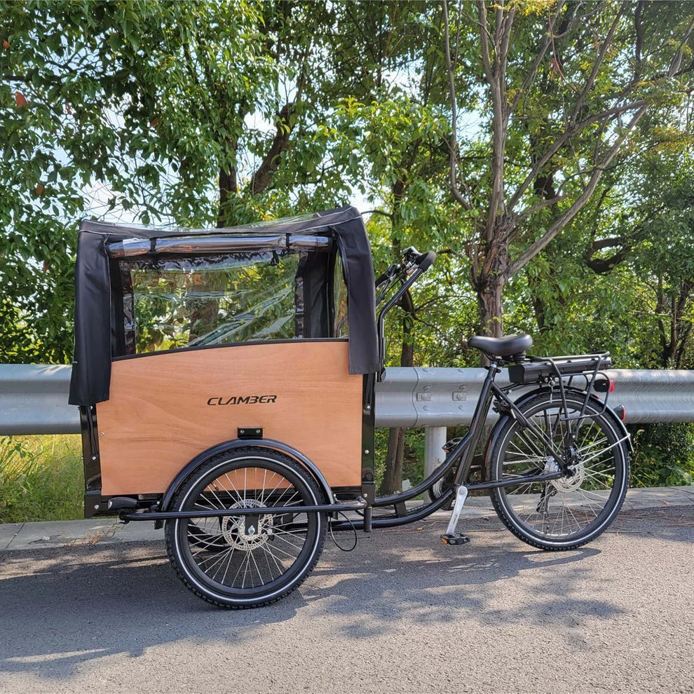 Trehjulig lådcykel med trälåda och svart regnkapell med fönster, parkerad på en asfaltväg vid ett vägräcke, gröna träd bakom
