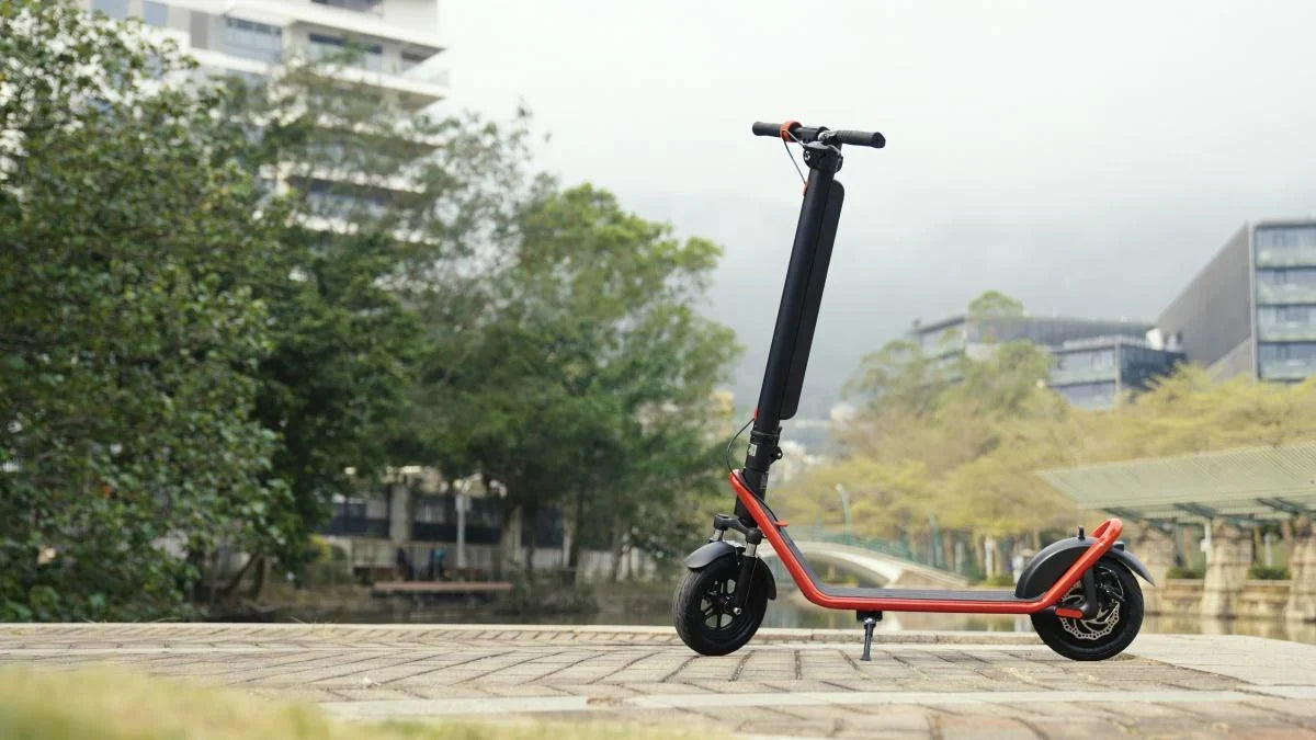 Svart elsparkcykel med röd ram från HX på stödbenet på en stenlagd promenad, med träd och moderna hus i bakgrunden