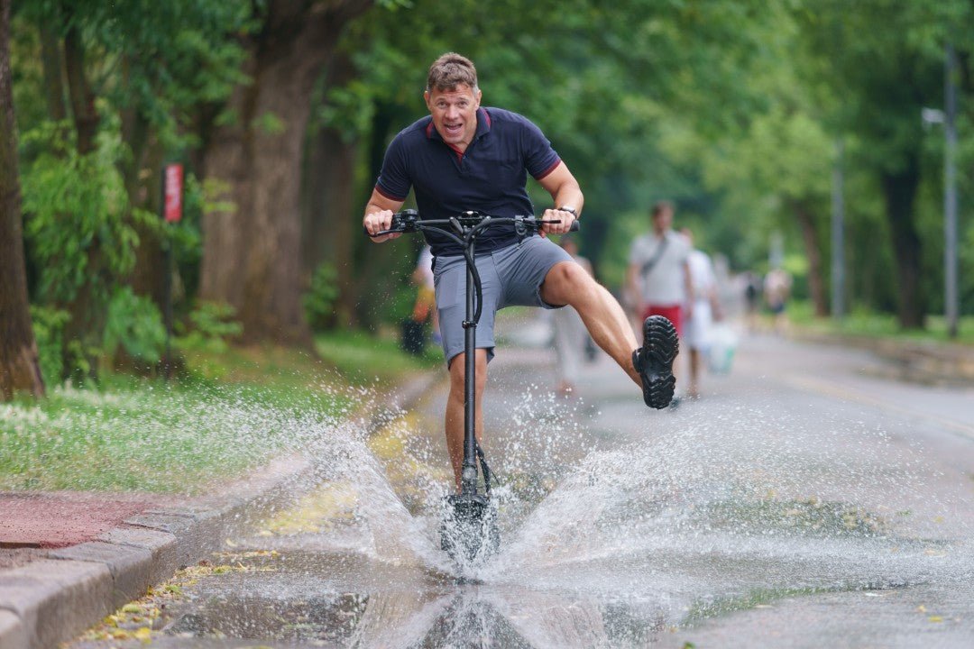 Man kör en elsparkcykel rakt genom en stor vattenpöl så att vattnet sprutar – körning i regn och IP-klassning