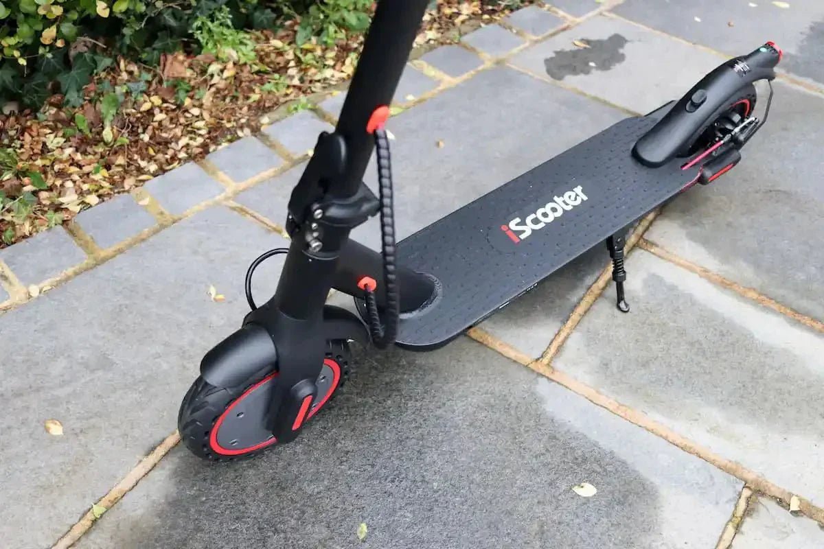 Svart iScooter-elsparkcykel med röda fälgdetaljer, uppställd på stödbenet på fuktiga grå stenplattor, höstlöv i kanten
