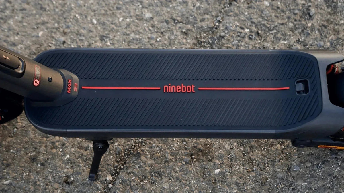 Ovanifrån på fotplattan till en svart Ninebot MAX G3 med röd ninebot-logga och utfällt stödben på grusig asfalt