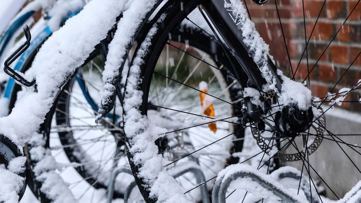 Snötäckta cyklar i ett cykelställ framför en tegelvägg – närbild på ett hjul med skivbroms medan snön faller