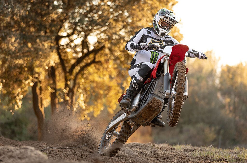 Crossförare i vit och svart utrustning gör en wheelie med en röd crossmotorcykel på en jordbana, träd i motljus bakom