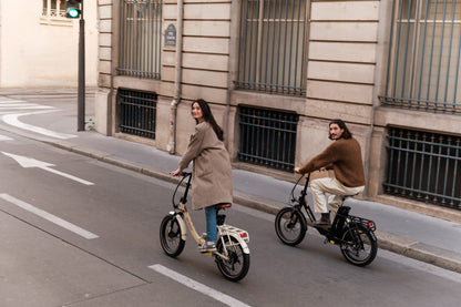 Två personer cyklar på en stadsgata på varsin hopfällbar cykel, en beige och en svart