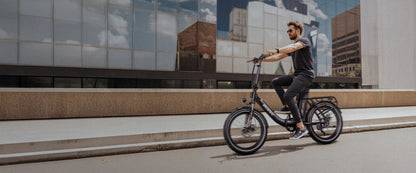 Man med solglasögon cyklar OneSport OT16 Pro längs en gata framför en glasfasad