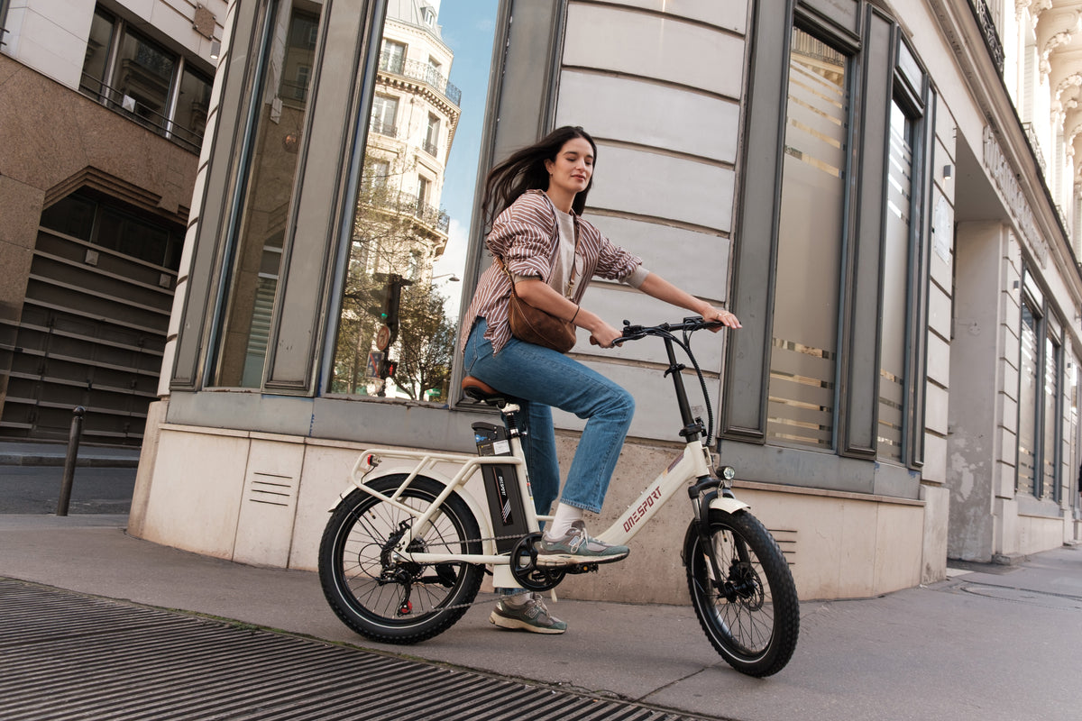 Kvinna cyklar OneSport OT16 Pro, vit elcykel, sedd från sidan på en stadsgata vid en stenbyggnad