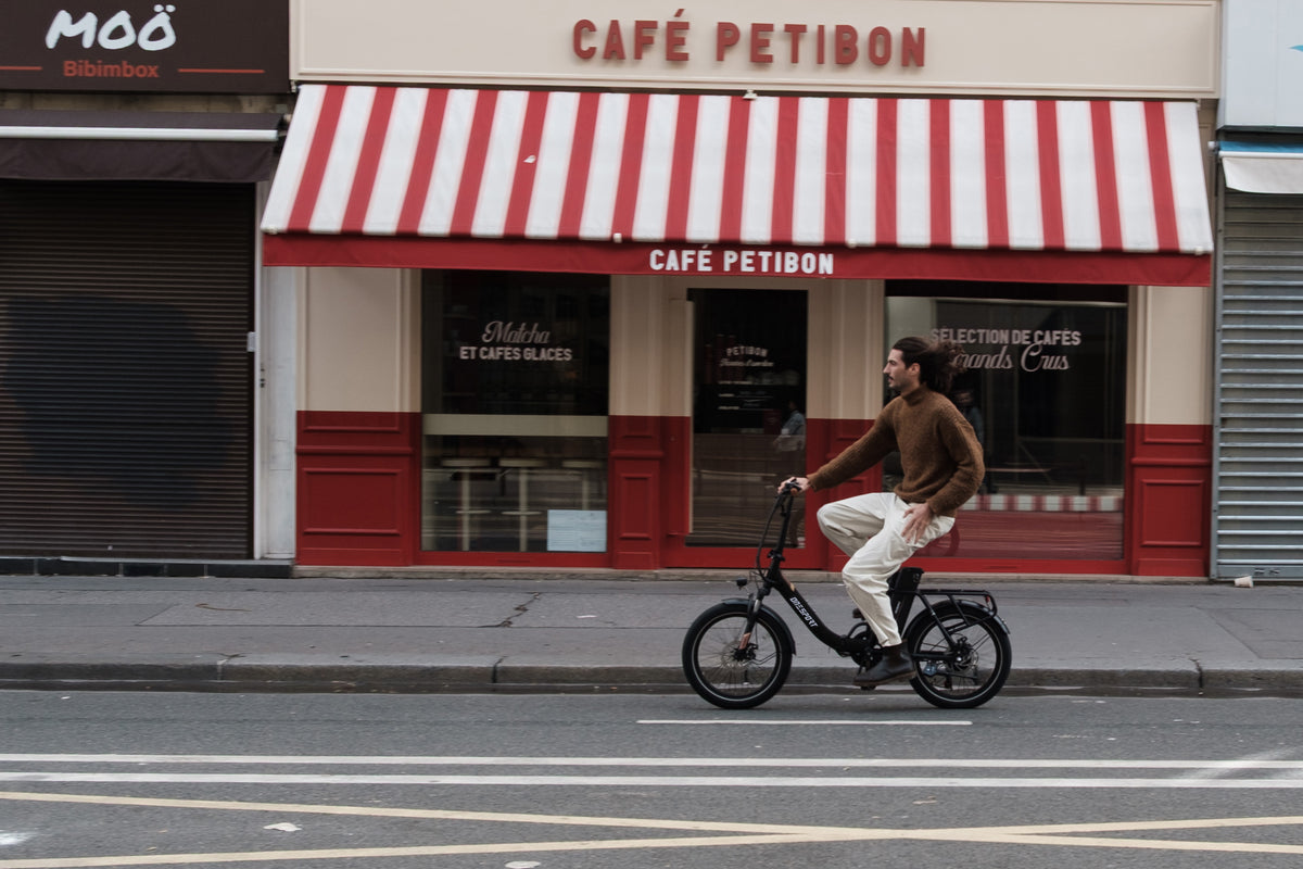 Man cyklar OneSport OT16 Pro, svart cykel, i sidovy framför ett kafé med rödvit randig markis