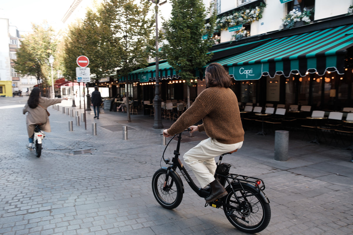 Person cyklar OneSport OT16 Pro, svart cykel, på kullerstensgata förbi ett kafé med grönrandig markis