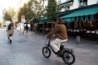 Person cyklar OneSport OT16 Pro, svart cykel, på kullerstensgata förbi ett kafé med grönrandig markis