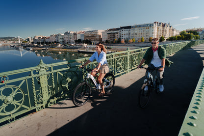 Två personer cyklar OneSport WT05 över en grön bro med flod och stad i bakgrunden