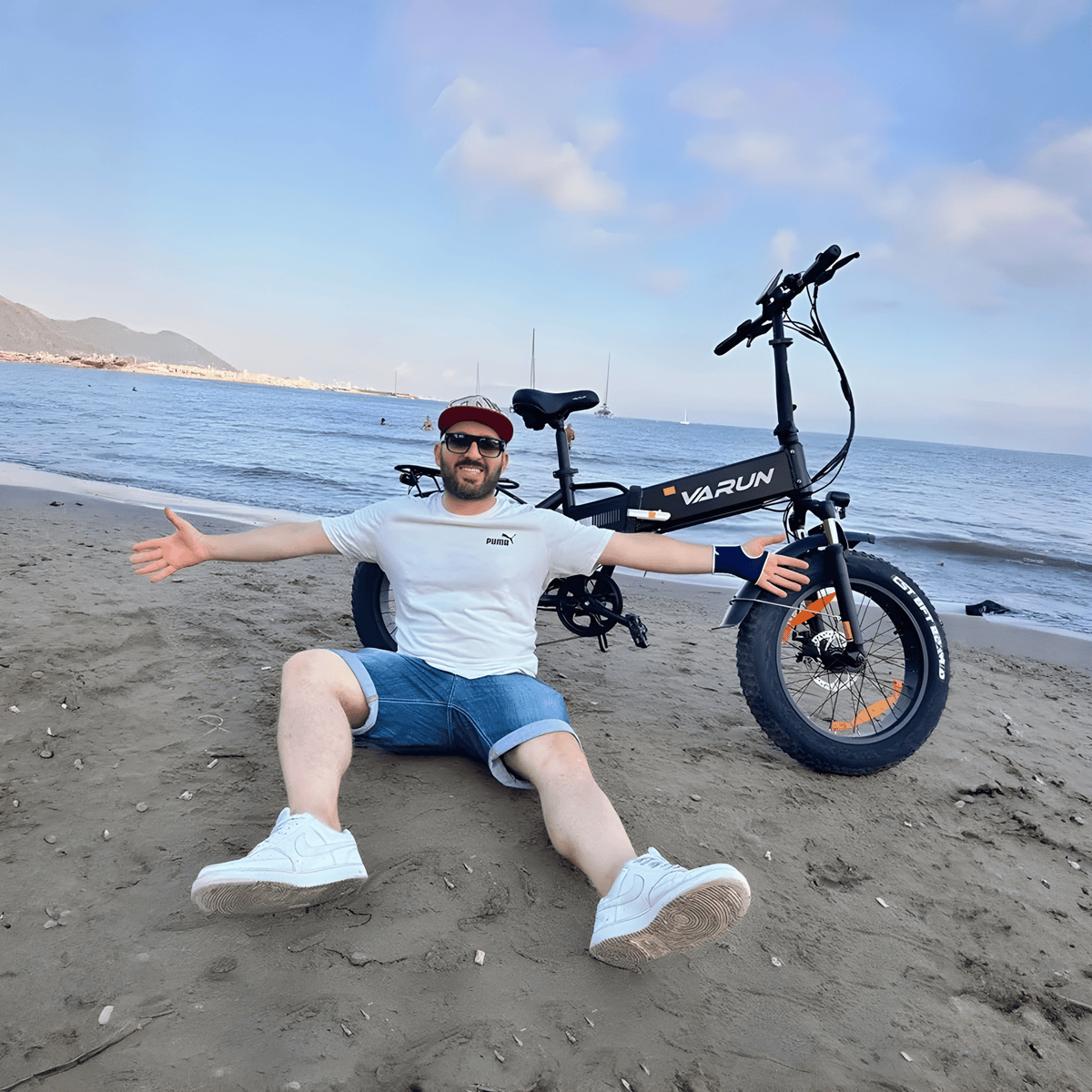Person sitter bredvid VARUN S20-1 hopfällbar fatbike-elcykel i färgen svart vid havet