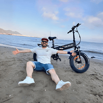 Person sitter bredvid VARUN S20-1 hopfällbar fatbike-elcykel i färgen svart vid havet